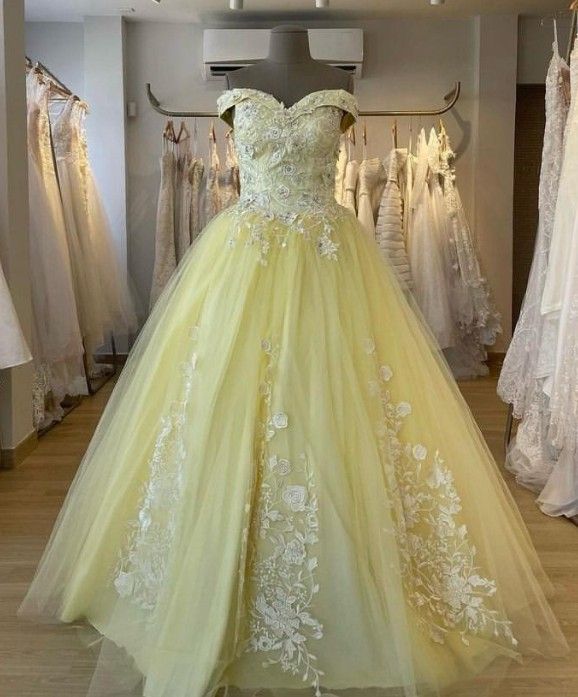 Vestido de novia amarillo de corte princesa con escote barco y apliques florales blancos, expuesto en una tienda nupcial.