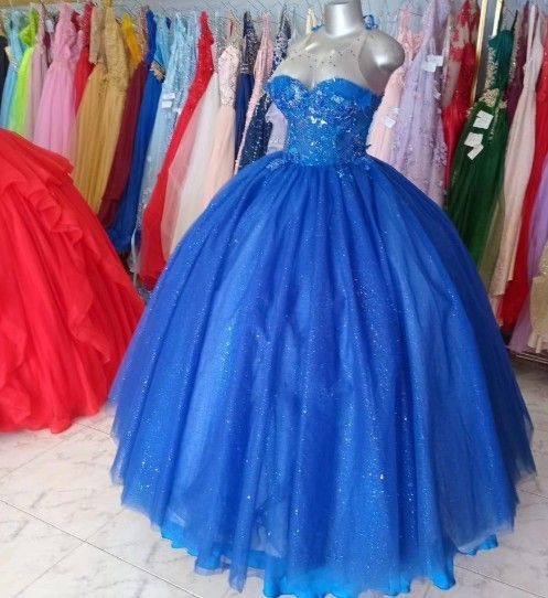 Vestido de gala azul sobre un maniquí, corpiño brillante, falda voluminosa, rodeado de otros vestidos coloridos.