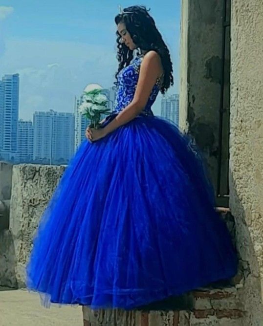 mujer con un vestido de gala azul, que sostiene flores blancas, está de pie en unas escaleras con el horizonte de la ciudad
