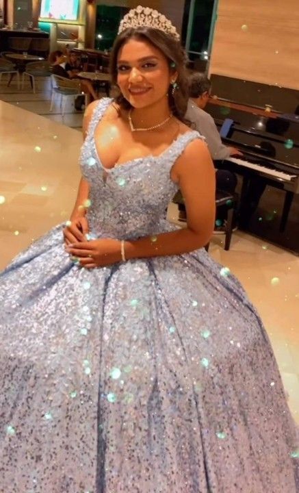 Una mujer con un vestido azul brillante y una tiara sonríe, sentada en una elegante habitación, con un piano al fondo.