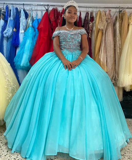Niña con un vestido de gala color verde azulado con corpiño plateado y corona, posando en una tienda de vestidos.