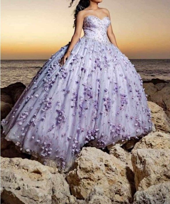 Mujer con vestido de gala palabra de honor color lavanda con adornos florales está de pie sobre unas rocas cerca del océano