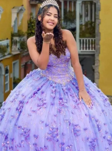 Mujer con vestido de gala color lavanda lanza un beso, luciendo una tiara. Escenario exterior con edificios.