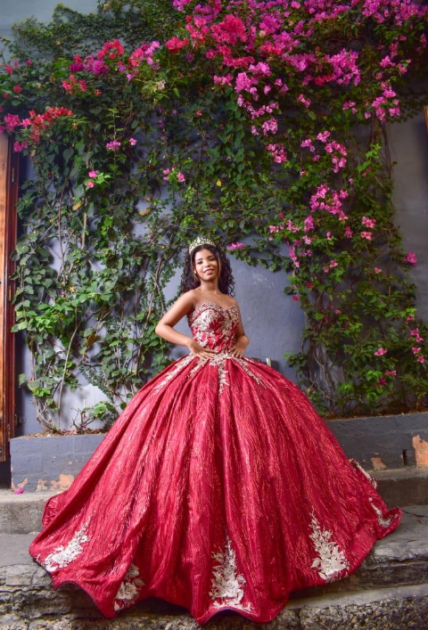 Mujer con un vestido de gala rojo con bordados dorados, posando frente a una pared de flores rosas y follaje verde.