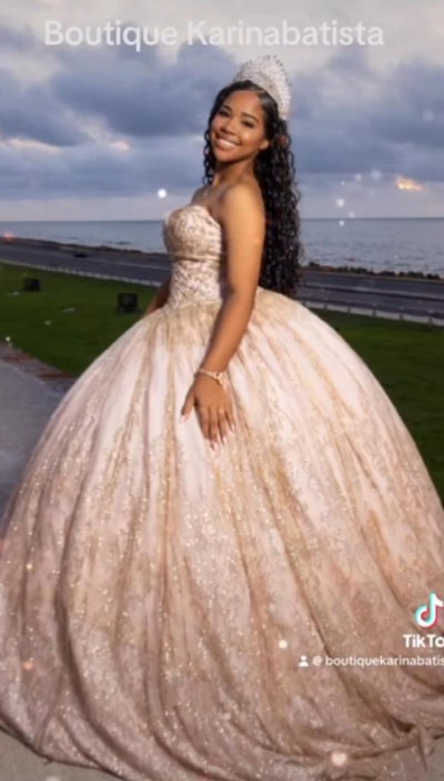 Una mujer con un vestido dorado sin tirantes posa al aire libre cerca del agua. Lleva una tiara y una pulsera.