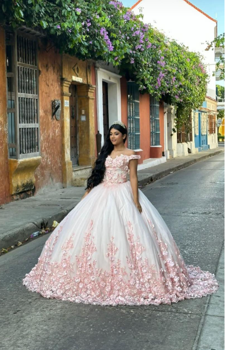 Una mujer con un vestido de gala rosa se encuentra de pie en una estrecha calle empedrada, bordeada de edificios coloridos.