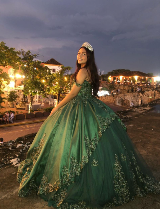 Joven con vestido de gala verde y tiara sonríe, posando al aire libre al atardecer.