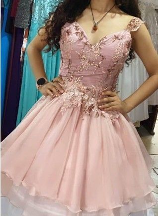 Mujer vestida con un vestido formal rosa claro con adornos florales y falda de capas, de pie en el interior.