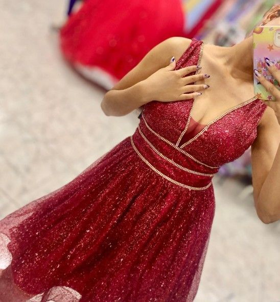 Mujer con un vestido rojo brillante de gala, en interiores, sosteniendo un teléfono; otros vestidos al fondo.