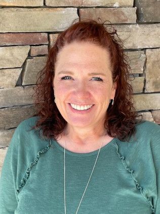 Woman with red hair, smiling at camera, wearing green shirt, against a stone wall.
