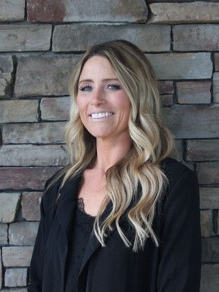 Woman with blonde hair smiles in front of a stone wall, wearing a black top and jacket.