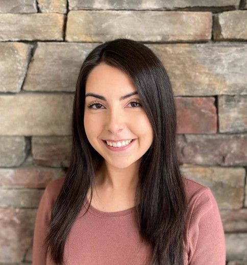 Woman with dark hair smiles, wearing a pink top, in front of a stone brick wall.