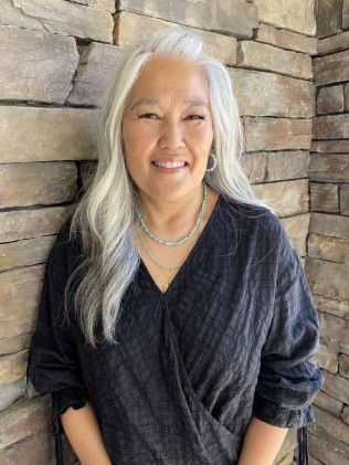 Woman with long gray hair smiles in front of a stone wall, wearing a dark top and necklace.