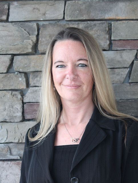 Woman with long blonde hair smiles, wearing a black blazer, in front of a stone wall.