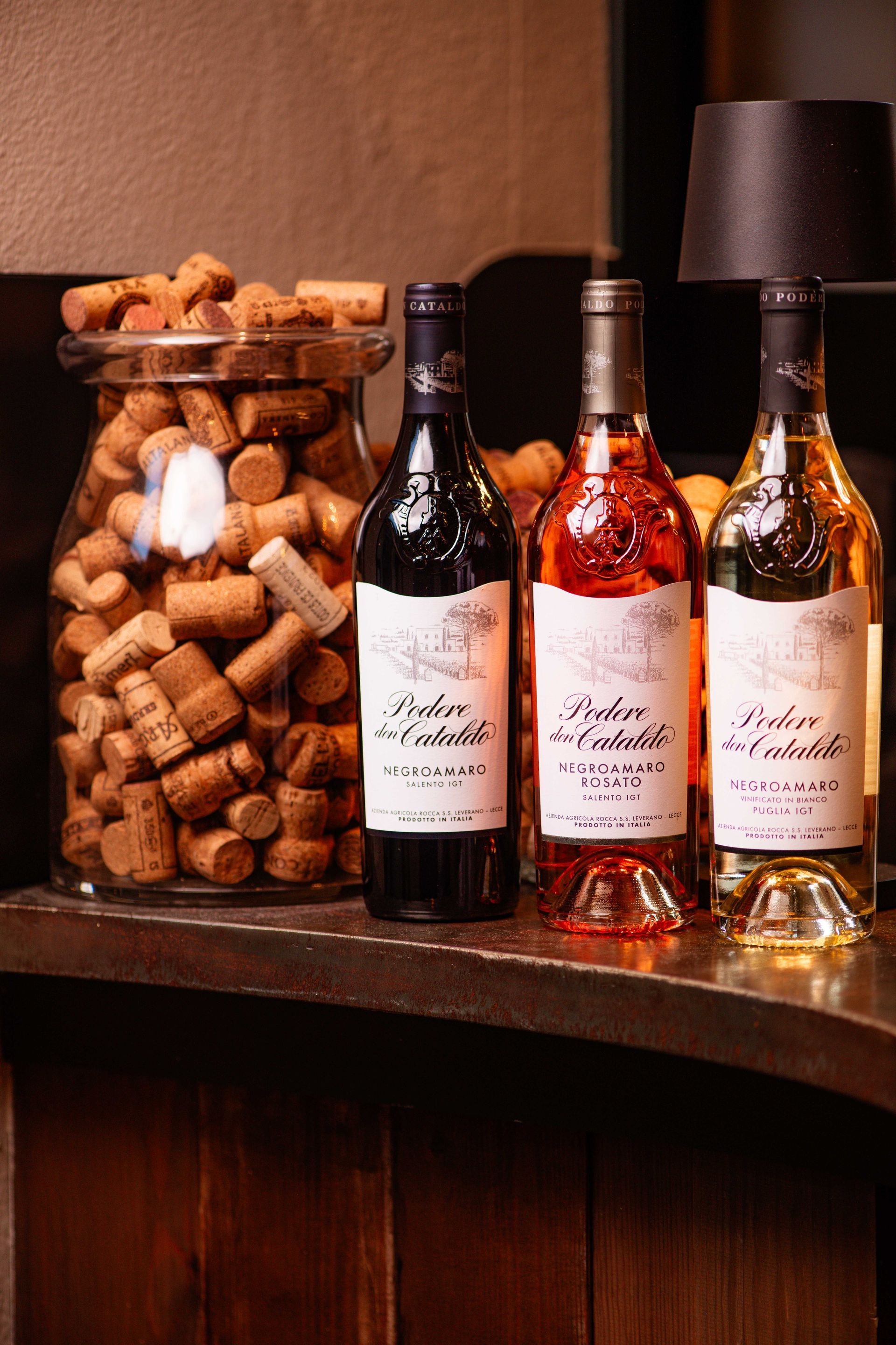 Tre bottiglie di vino sono appoggiate su un tavolo accanto a un barattolo di tappi di sughero.