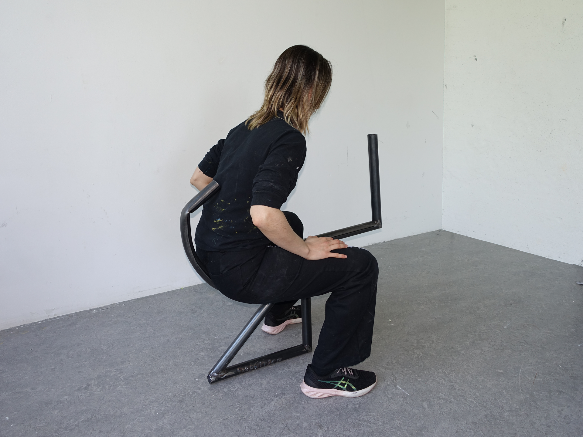 Woman in black clothing sits on a minimalist metal chair in a white room, looking away.
