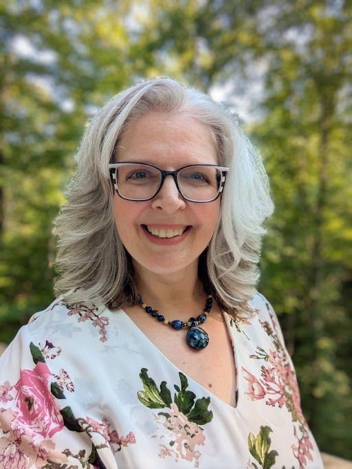 Woman with gray hair and glasses smiles outdoors, wearing floral top and necklace.