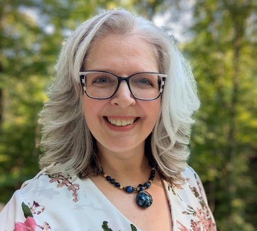 Woman with gray hair, glasses, smiling, wearing floral top, necklace, outdoors.