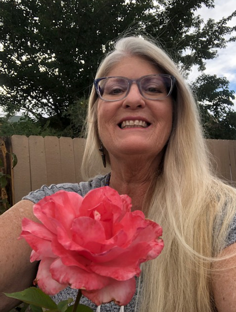 A woman wearing glasses is holding a pink rose and smiling.