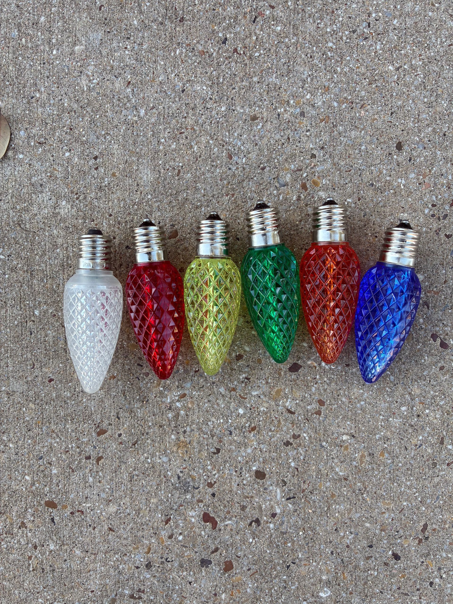 Six colorful Christmas light bulbs arranged on a gray concrete surface.