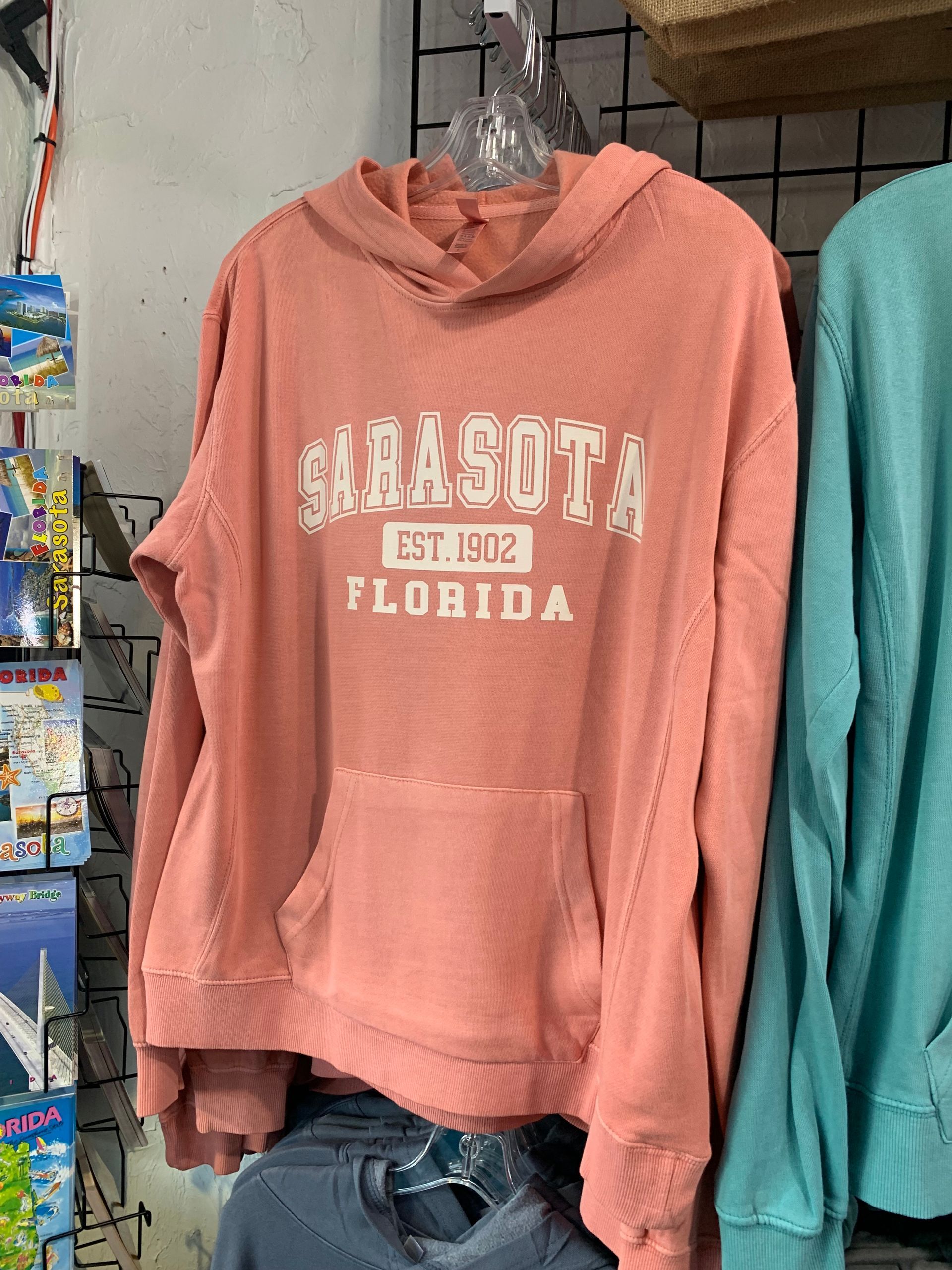 Pink Sarasota hoodie on a rack, with white lettering and a front pocket, in a shop setting.