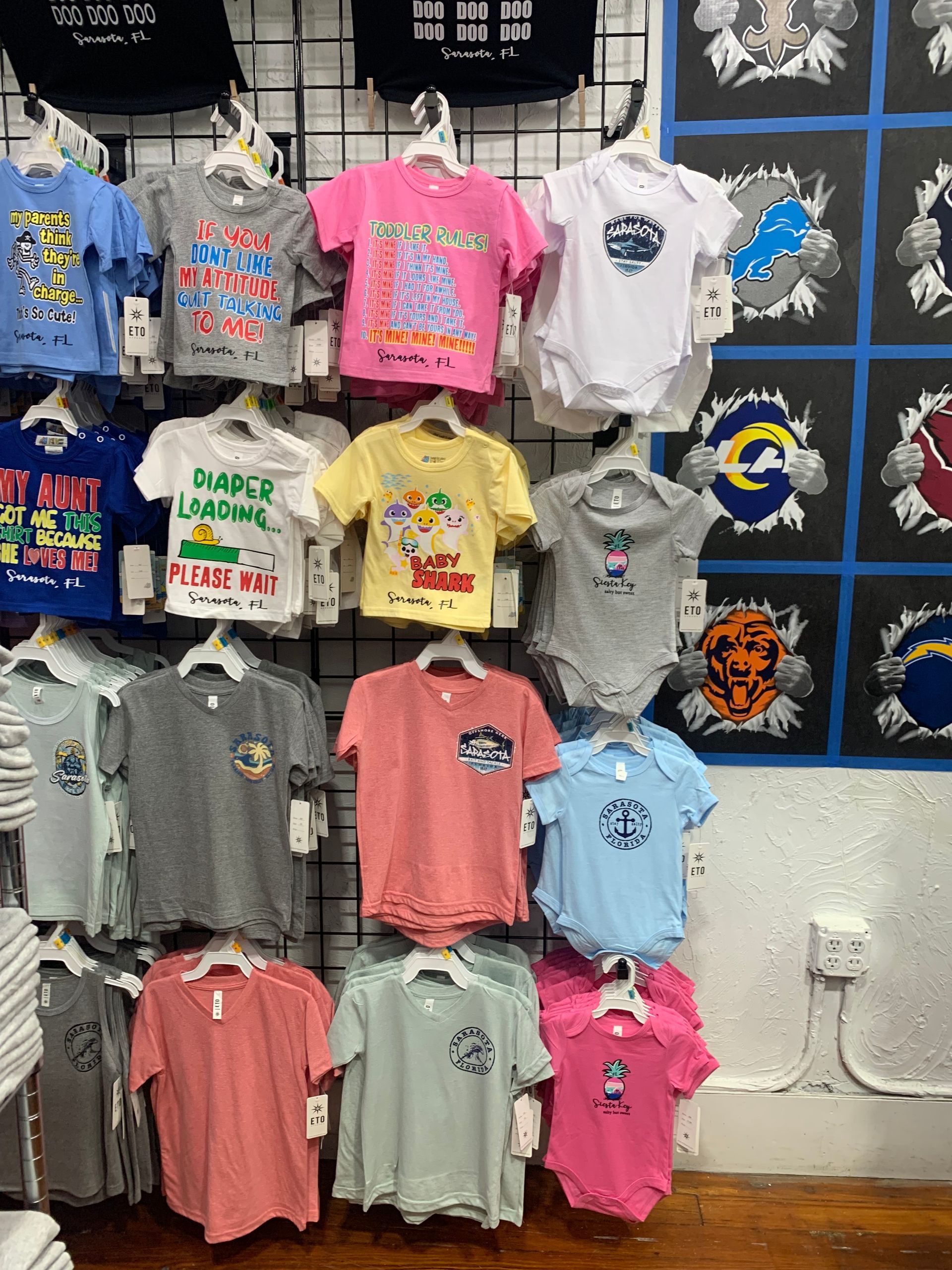 T-shirts and baby onesies in various colors hanging on a retail display. Some have graphic designs.