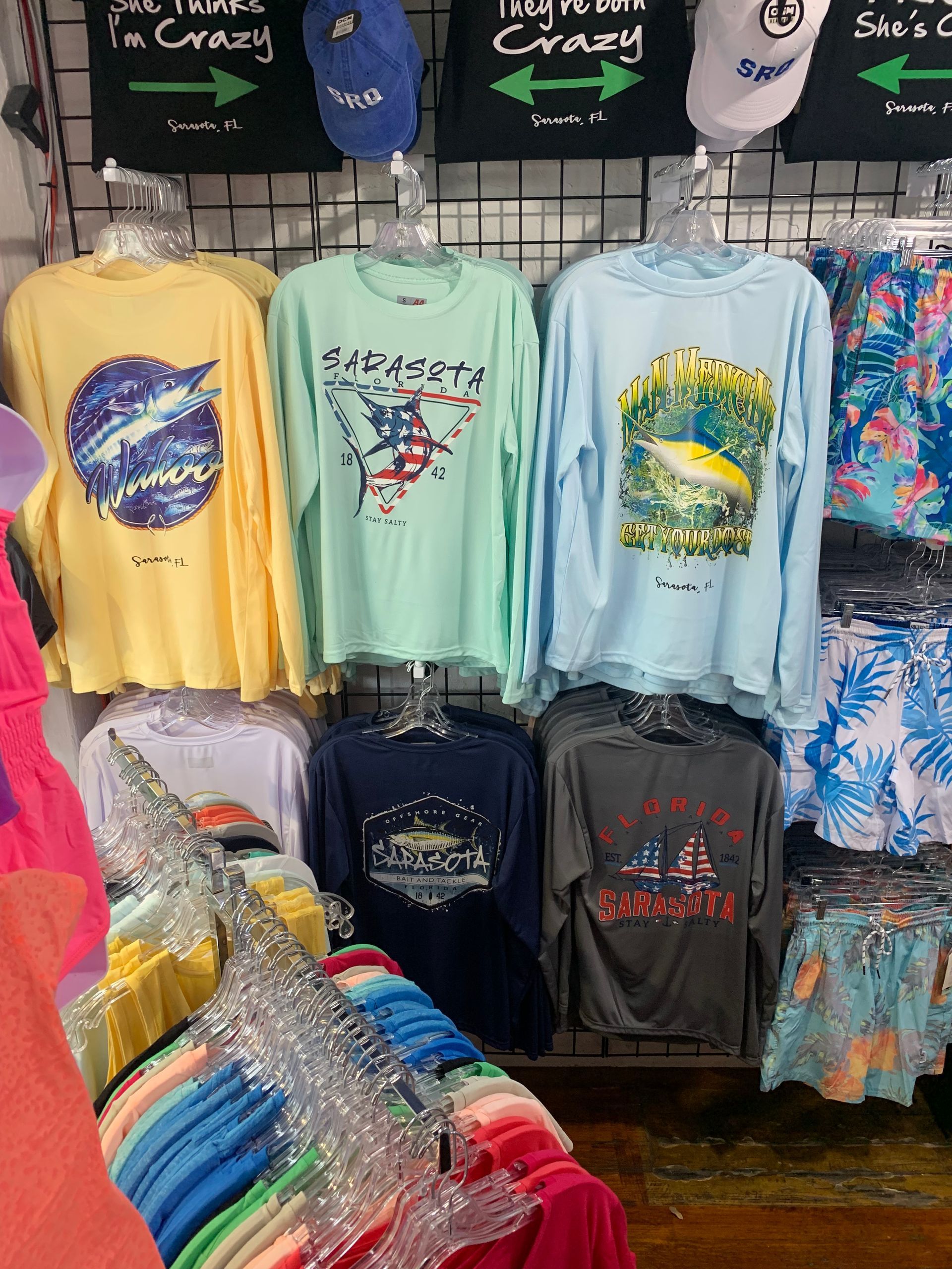 Shirts with various designs on display in a store; hats and colorful shorts are also visible.