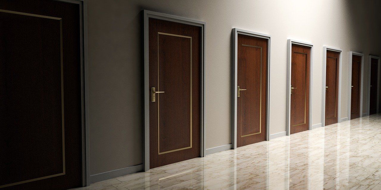 A hallway with several closed brown doors, some with gold trim, a dark floor, and bright light.