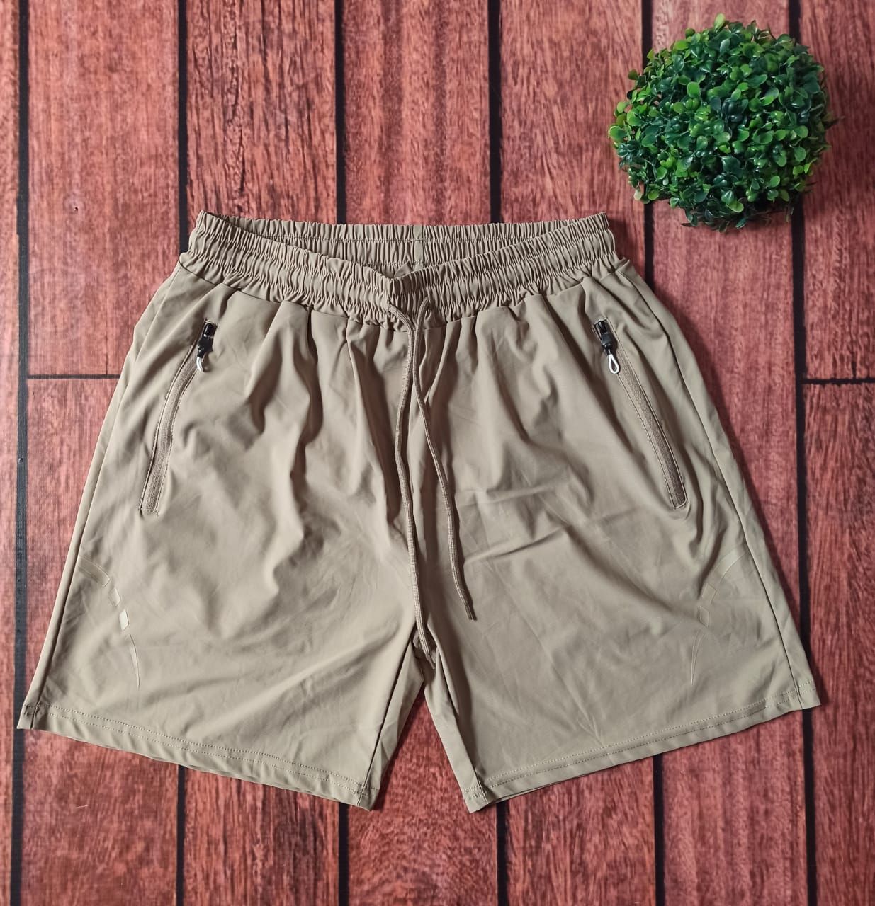 Pantalón corto deportivo beige con cordón, dos bolsillos con cremallera, sobre fondo de madera con una pequeña planta verde.
