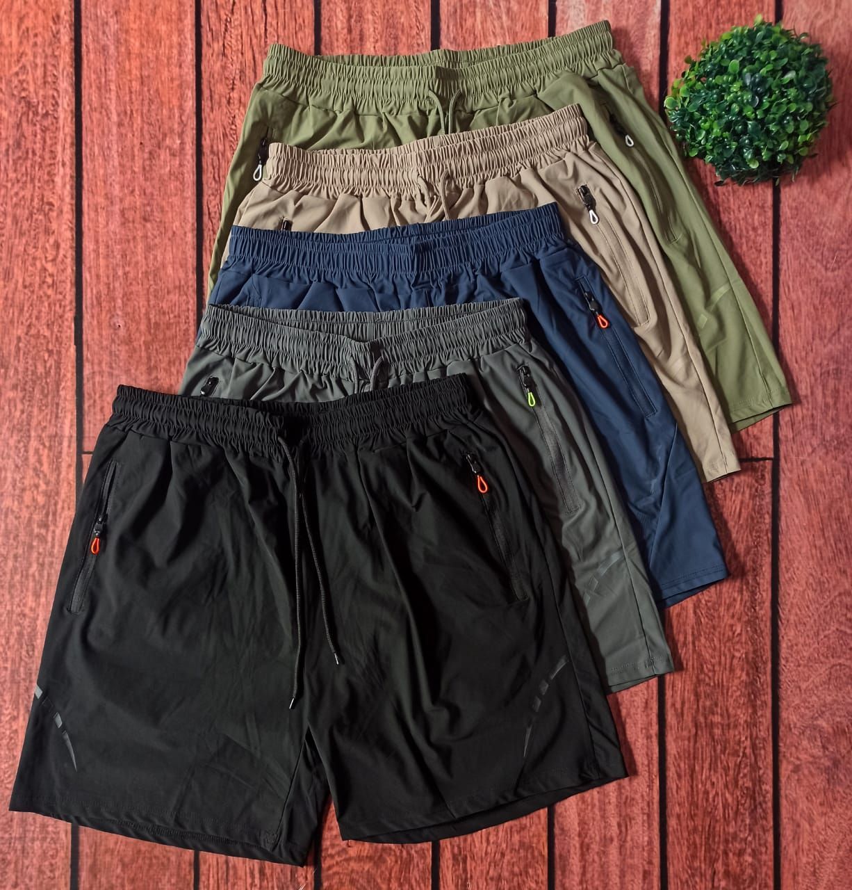 Pantalón corto deportivo beige con cordón, dos bolsillos con cremallera, sobre fondo de madera con una pequeña planta verde.