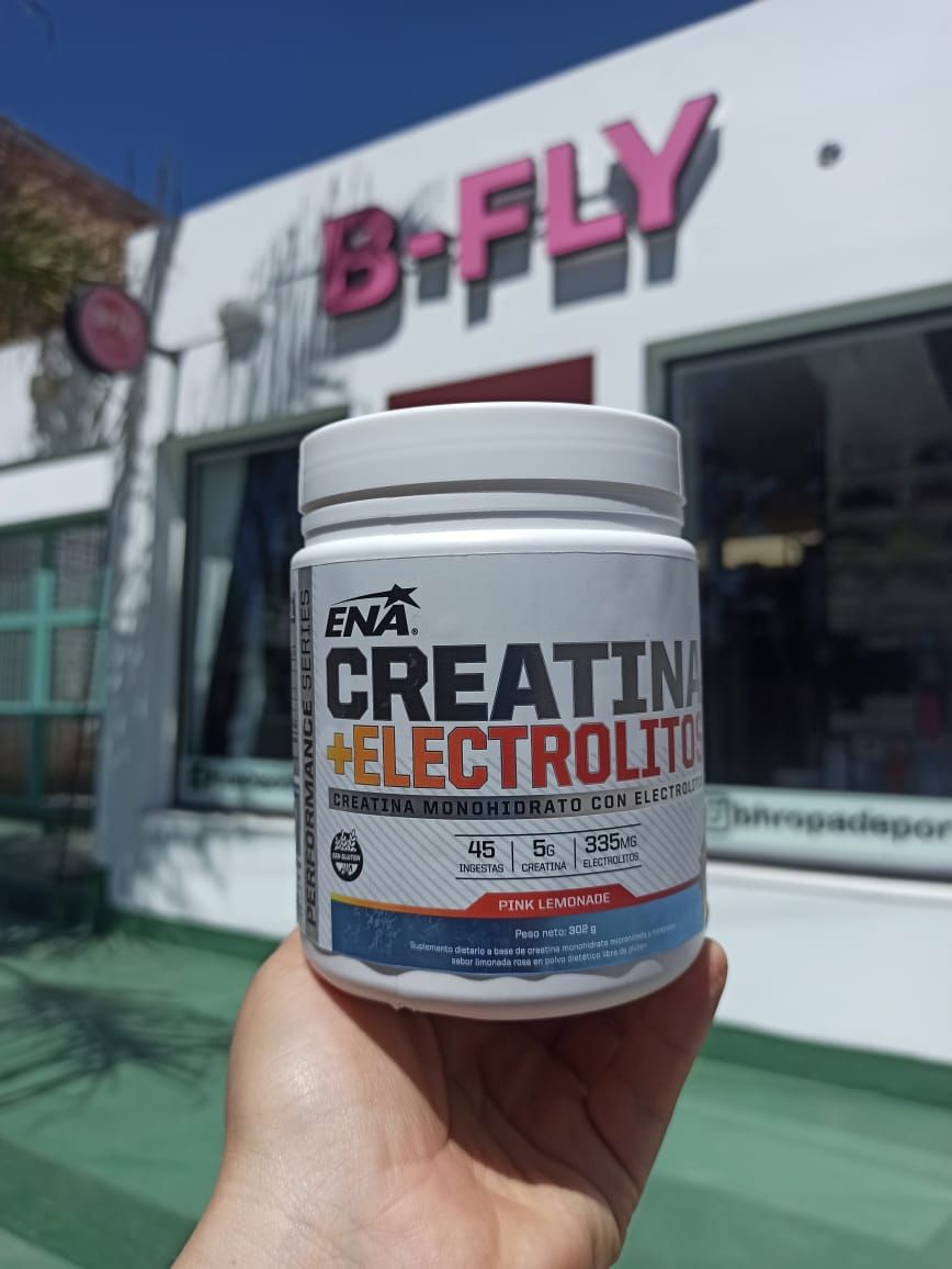Mano sosteniendo un envase de suplemento etiquetado "Creatina + Electrolitos" frente a una tienda "B-FLY".