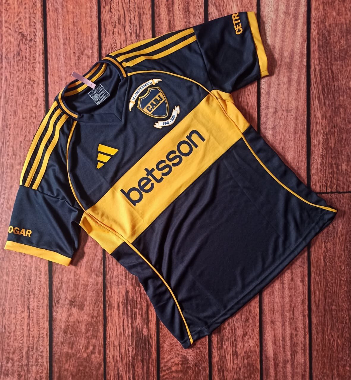 Camiseta de fútbol azul oscuro y dorado con el logotipo de Adidas y el patrocinador "betsson" sobre un fondo de madera.