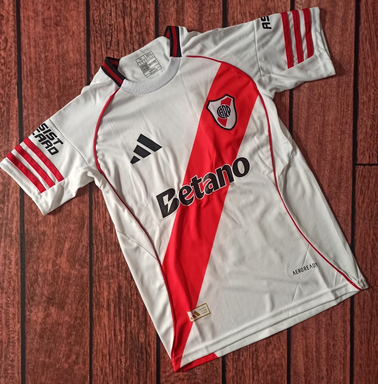 Camiseta de fútbol blanca con franja y acentos rojos; logotipo de Betano, logotipo de Adidas, escudo de River Plate.
