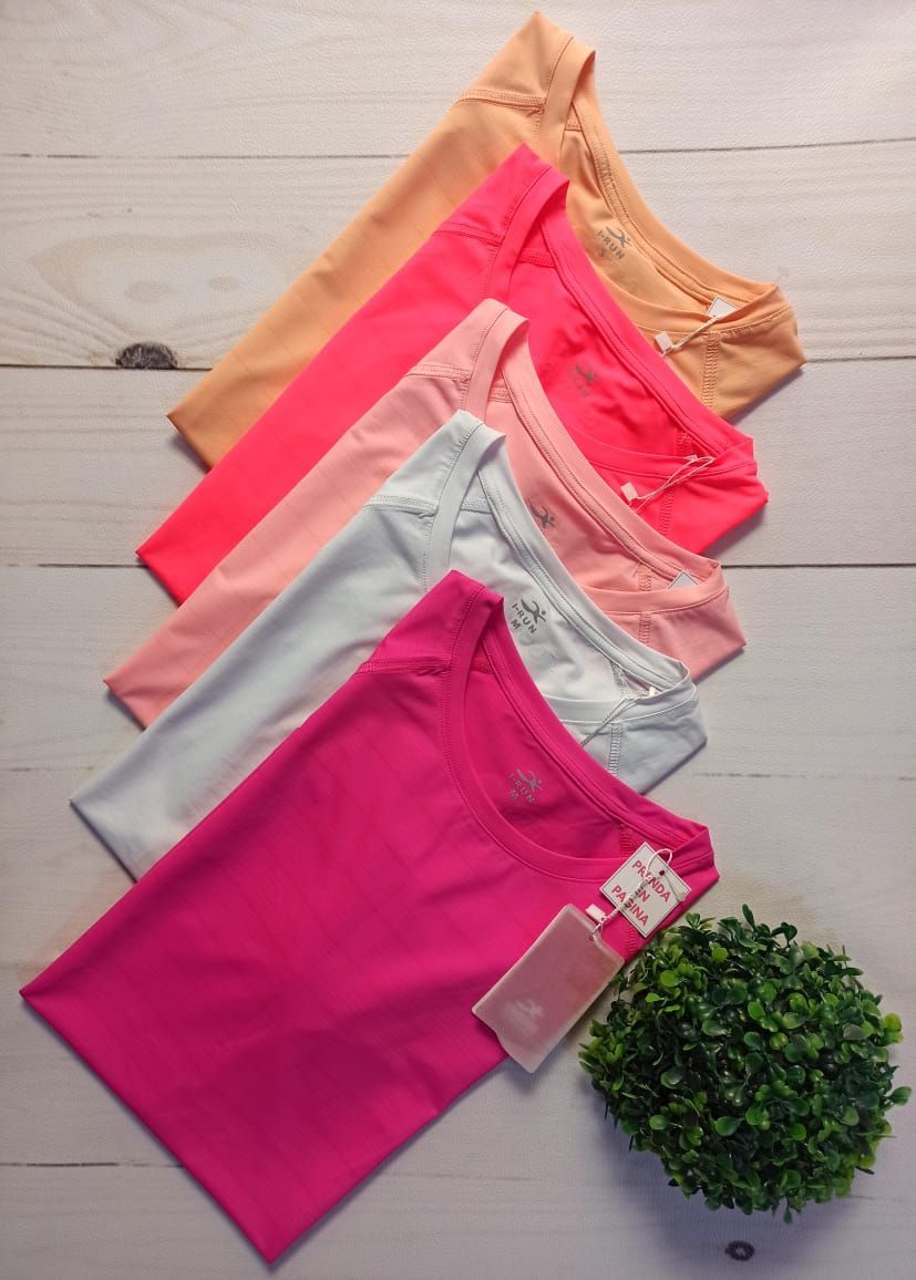 Camisetas de colores brillantes apiladas sobre una superficie de madera blanca, con un pequeño arbusto al costado.