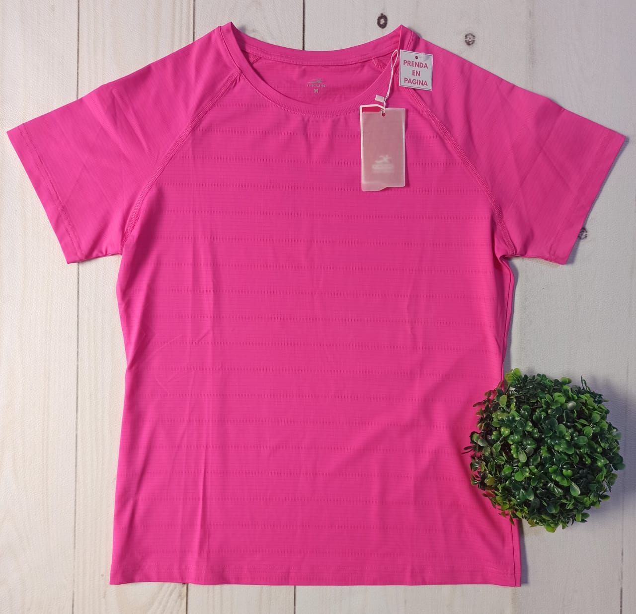 Camisetas de colores brillantes apiladas sobre una superficie de madera blanca, con un pequeño arbusto al costado.
