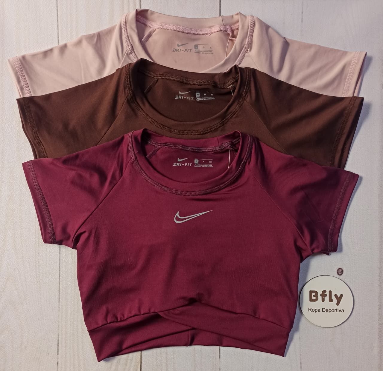 Tres crop tops de Nike, de color rosa, marrón y burdeos, sobre una superficie de madera blanca.
