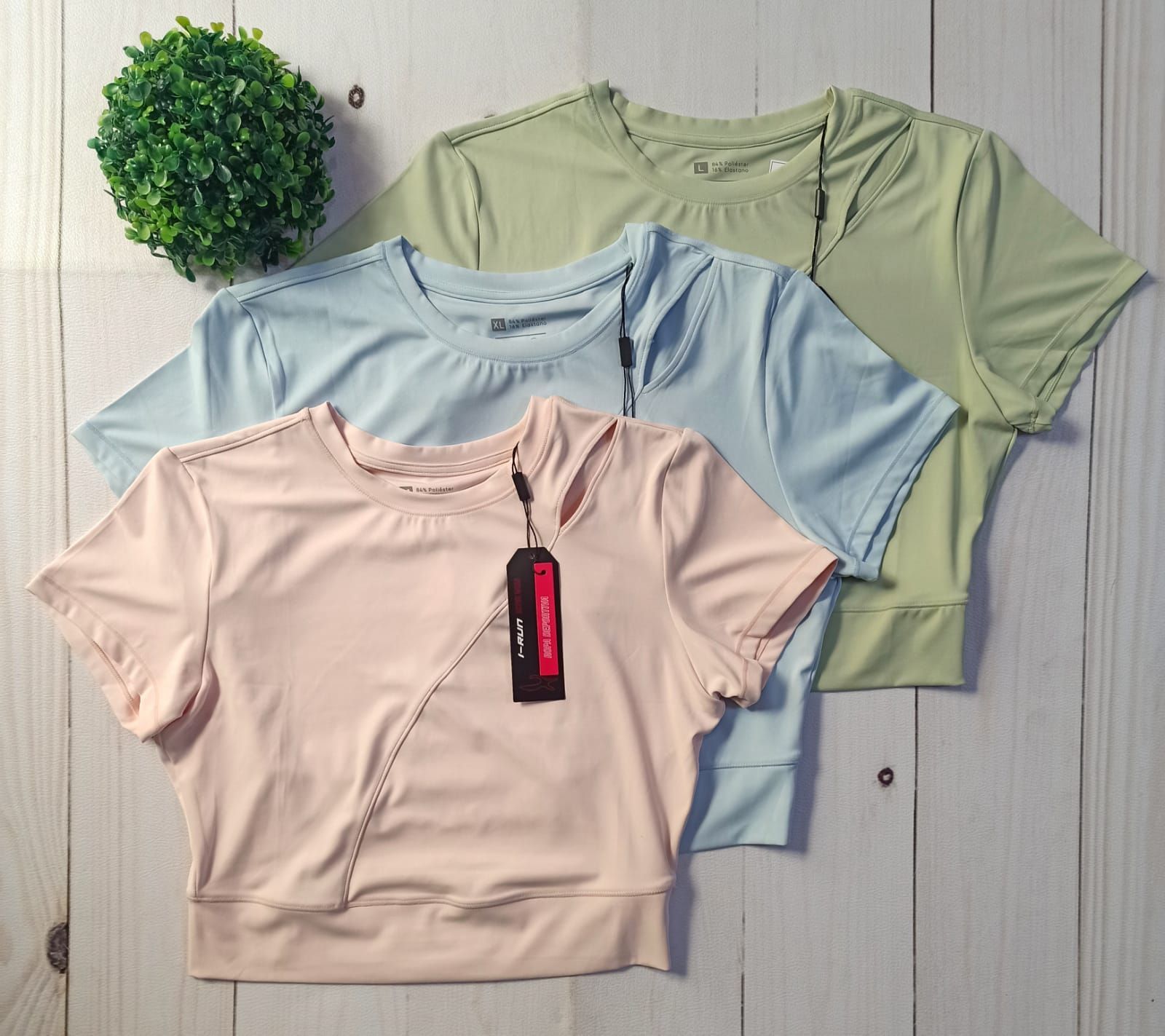 Tres camisetas deportivas de manga corta: melocotón, azul y verde; dispuestas sobre una superficie con vetas de madera.