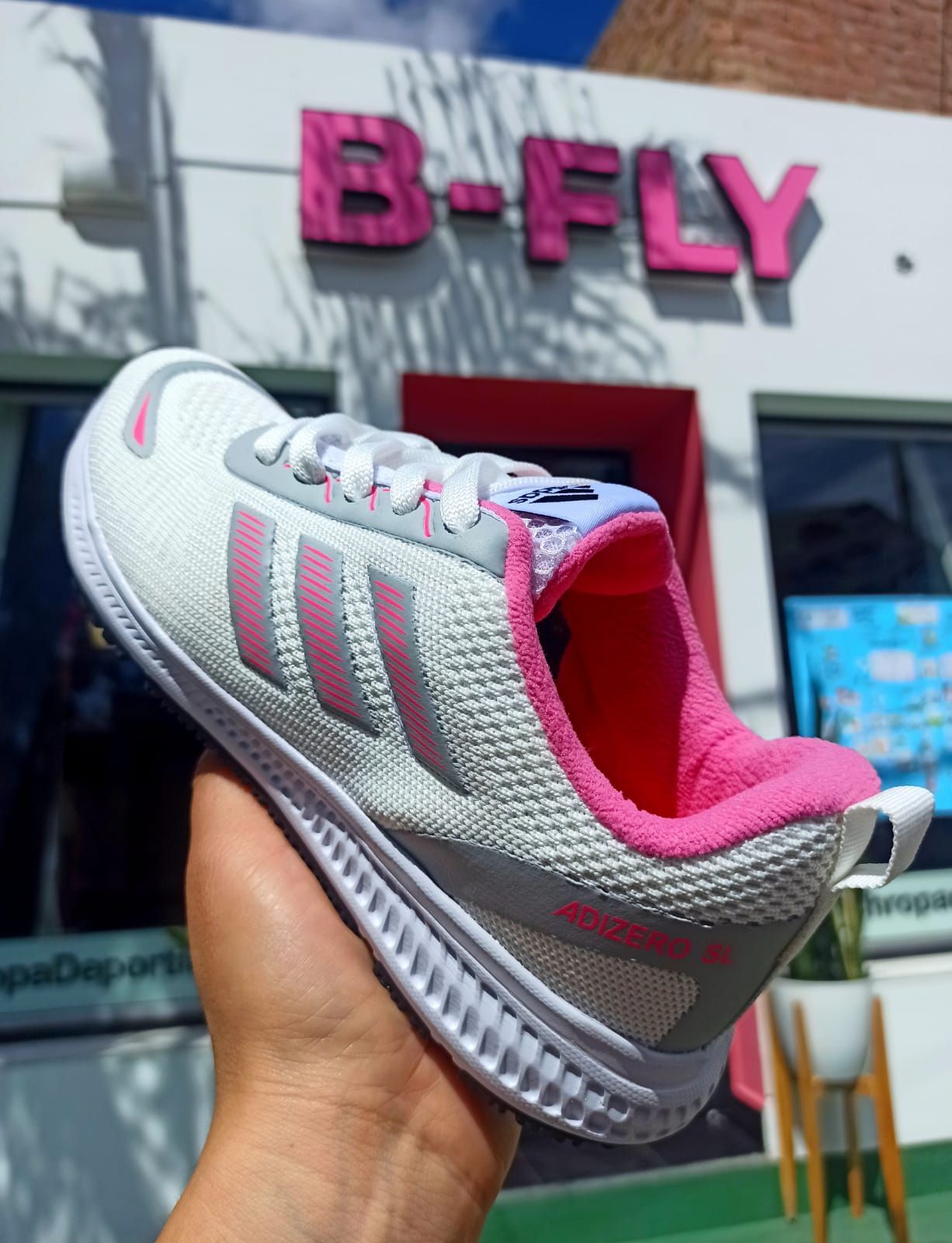 Zapatilla deportiva Adidas blanca con detalles en rosa, colocada frente a la fachada de una tienda.