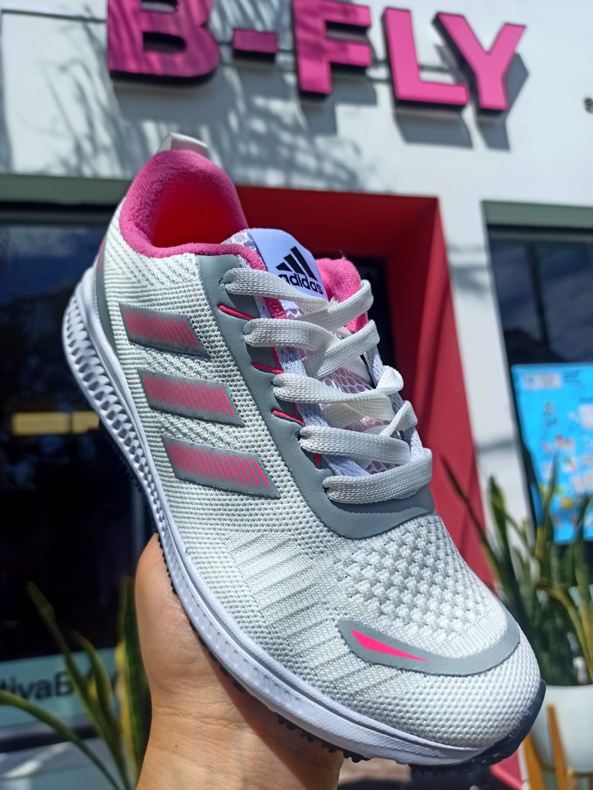 Zapatilla deportiva Adidas blanca con detalles en rosa, colocada frente a la fachada de una tienda.