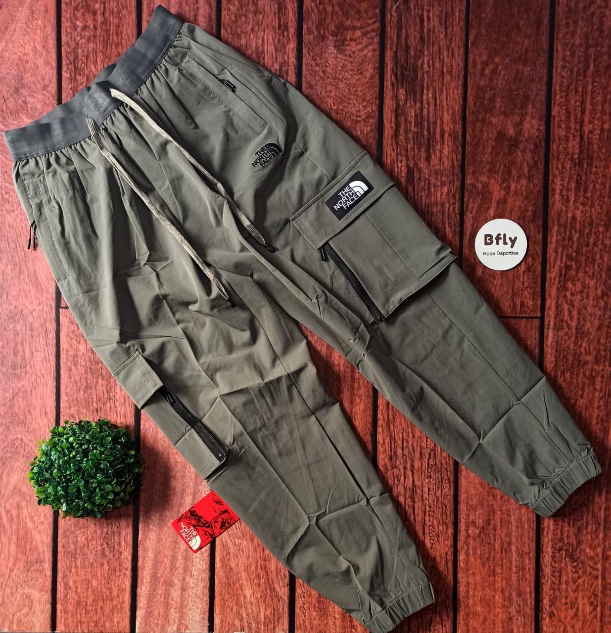 Tres pares de pantalones cargo, marrón, negro y gris, apilados sobre una superficie de madera con una pequeña planta verde.