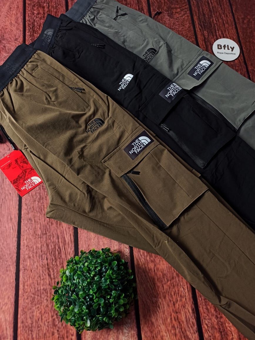Tres pares de pantalones cargo, marrón, negro y gris, apilados sobre una superficie de madera con una pequeña planta verde.