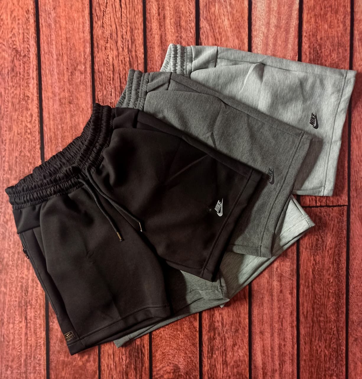 Tres pares de pantalones cortos Nike en negro, gris y gris claro apilados sobre una superficie de madera.