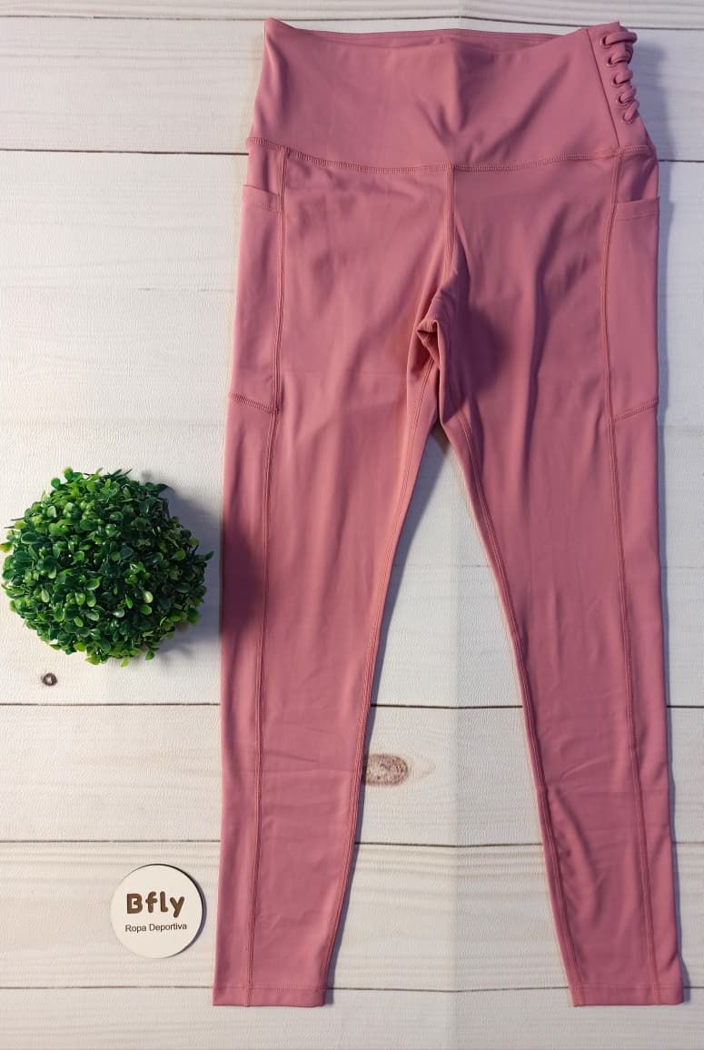Leggings rosas de cintura alta con bolsillos laterales, sobre un fondo de madera junto a una planta verde.