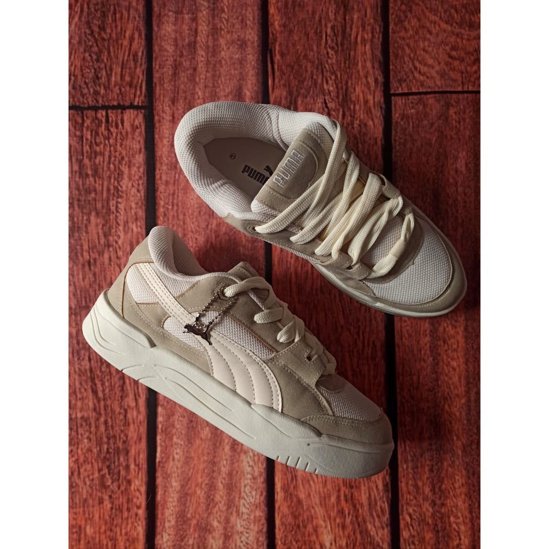 Un par de zapatillas deportivas color beige sobre un suelo de madera rojiza, vistas desde arriba.