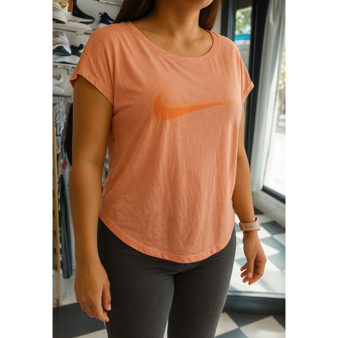 Mujer con camiseta Nike coral y leggings grises. Logotipo de Nike visible. Interior, junto a una ventana.