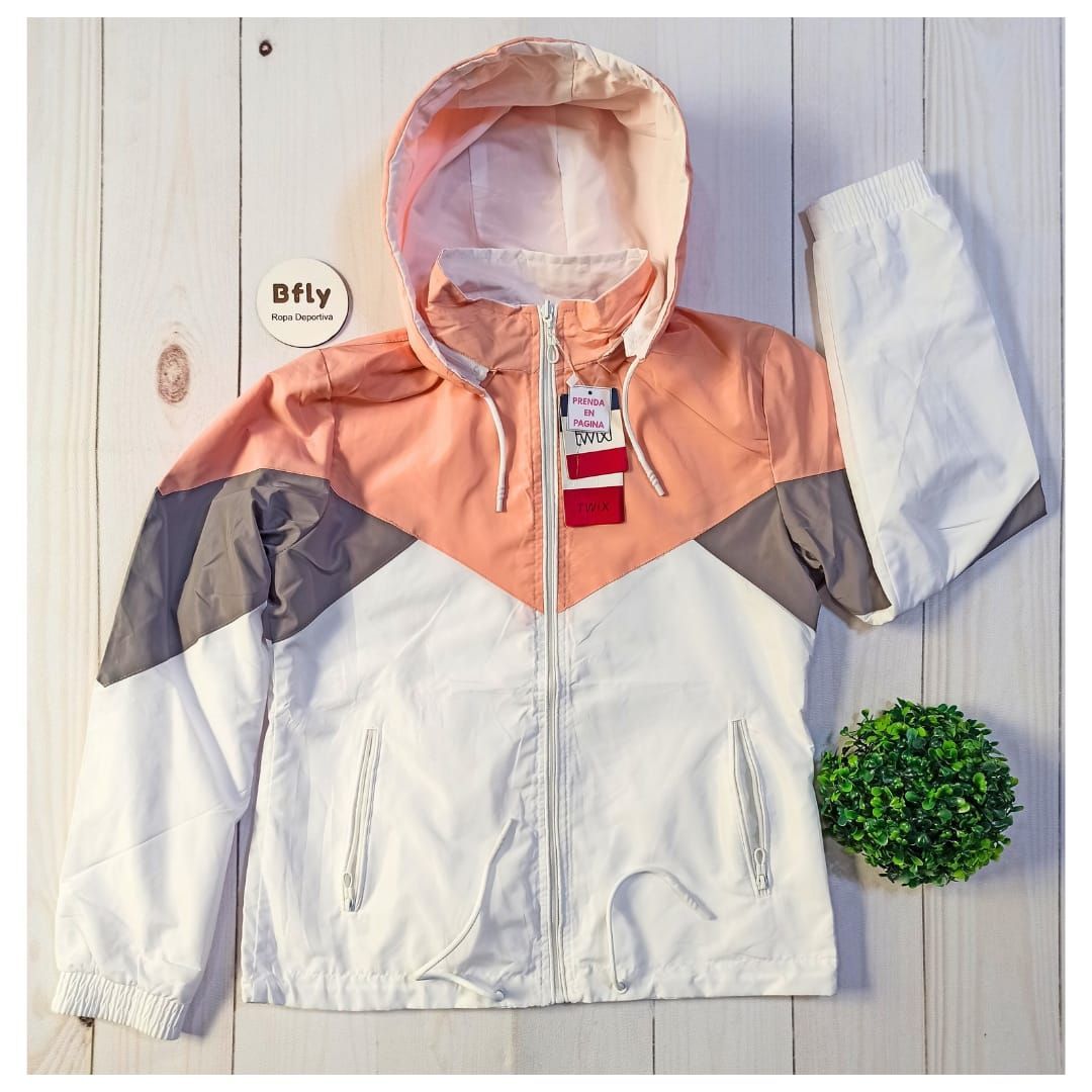 Mujer con una chaqueta con capucha, cremallera y bolsillos de color salmón, blanca y gris, de pie al aire libre.