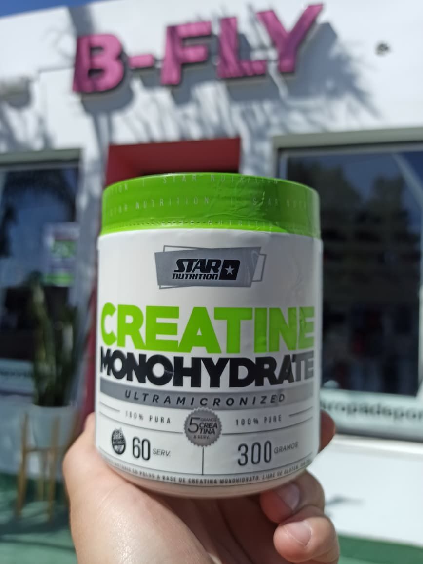 Mano sosteniendo un recipiente de monohidrato de creatina de Star Nutrition con tapa verde, frente a un edificio.