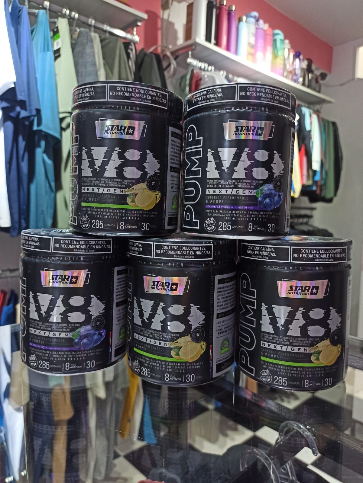 Cinco botes negros de bebida energética V8 apilados en un pasillo de una tienda, con ropa y botellas colgadas al fondo.