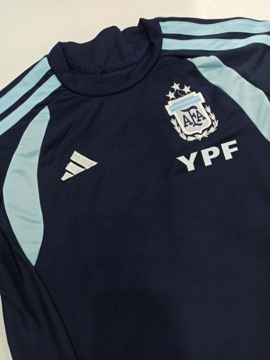 Camiseta de la selección argentina de fútbol, ​​azul marino oscuro con mangas azul claro y el logotipo del patrocinador YPF, colocada sobre una superficie plana.