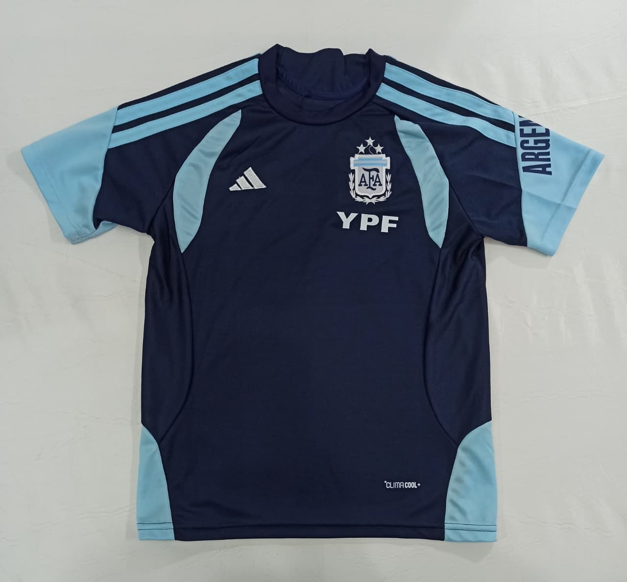Camiseta de la selección argentina de fútbol, ​​azul marino oscuro con mangas azul claro y el logotipo del patrocinador YPF, colocada sobre una superficie plana.